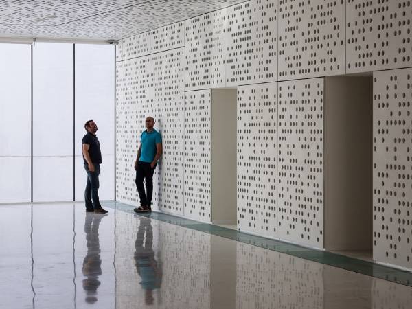 2 men are chatting inside the room fitted with white perforated metal sheets